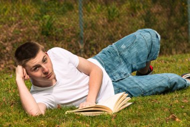 Genç bir çocuk halka açık bir parkta güneşin altında kitap okuyor. Beyaz tişört ve mavi kot pantolon giyiyor.
