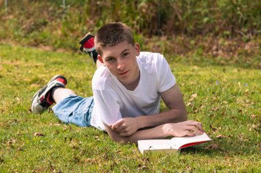 Genç bir çocuk halka açık bir parkta güneşin altında kitap okuyor. Beyaz tişört ve mavi kot pantolon giyiyor.