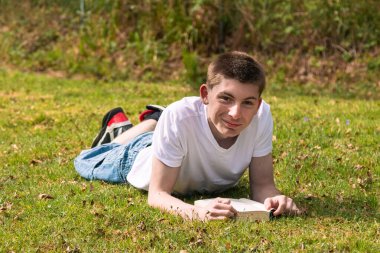 Mutlu ergen çocuk halka açık bir parkta güneşin altında kitap okuyor. Beyaz tişört ve mavi kot pantolon giyiyor.