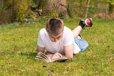 Genç bir çocuk halka açık bir parkta güneşin altında kitap okuyor. Beyaz tişört ve mavi kot pantolon giyiyor.