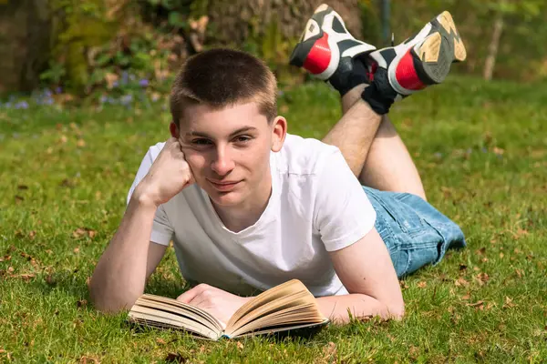 Genç bir çocuk halka açık bir parkta güneşin altında kitap okuyor. Beyaz tişört ve mavi kot pantolon giyiyor.