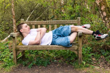 Halka açık bir parkta bankta beyaz tişört ve mavi kot pantolon giymiş genç bir çocuk uyuyor.