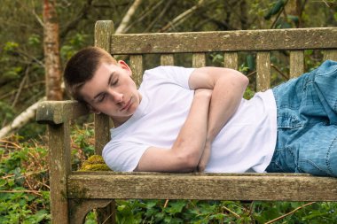 Halka açık bir parkta bankta beyaz tişört ve mavi kot pantolon giymiş genç bir çocuk uyuyor.