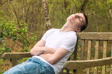 Halka açık bir parkta bankta beyaz tişört ve mavi kot pantolon giymiş genç bir çocuk uyuyor.