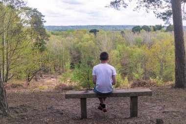 Genç bir çocuk parktaki bankta tek başına oturuyor manzaraya bakıyor, beyaz tişört ve mavi kot pantolon giyiyor.