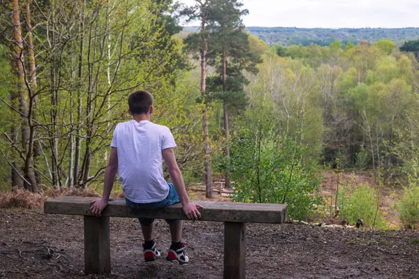 Genç bir çocuk parktaki bankta tek başına oturuyor manzaraya bakıyor, beyaz tişört ve mavi kot pantolon giyiyor.