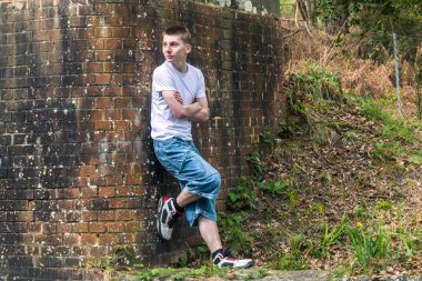 Tuğla duvara dayanmış beyaz tişört ve mavi kot giyen genç bir çocuk.