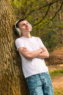 Beyaz t-shirt ve mavi kot giyen genç bir çocuk, halka açık bir parkta, bir ağaca yaslanmış kulaklıkla müzik dinliyor.