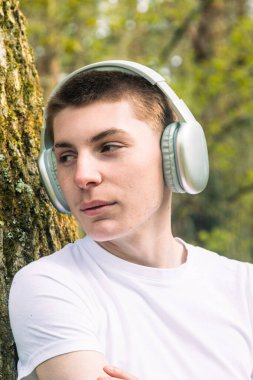 Beyaz t-shirt ve mavi kot giyen genç bir çocuk, halka açık bir parkta, bir ağaca yaslanmış kulaklıkla müzik dinliyor.