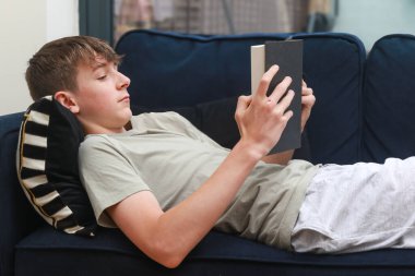 Kanepeye uzanmış roman okuyan bir genç çocuk.