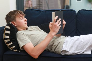 Kanepeye uzanmış roman okuyan bir genç çocuk.