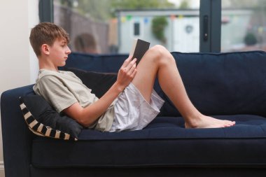 Kanepeye uzanmış roman okuyan bir genç çocuk.