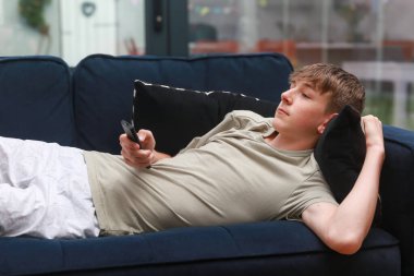 Kanepede uzanmış uzaktan kumandayla televizyon seyreden bir genç.