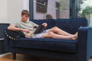 Koltukta uzanmış, bir kase abur cuburla roman okuyan genç bir çocuk.