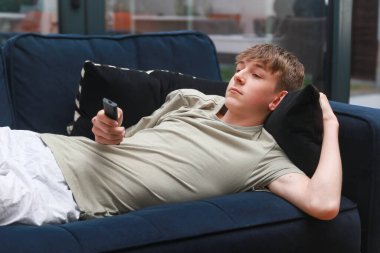 Kanepede uzanmış uzaktan kumandayla televizyon seyreden bir genç.