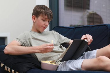 Koltukta uzanmış, bir kase abur cuburla roman okuyan genç bir çocuk.