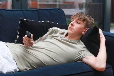 Kanepede uzanmış uzaktan kumandayla televizyon seyreden bir genç.