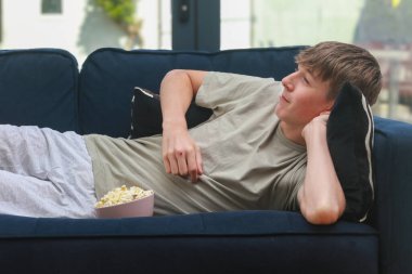 Televizyon izlerken elinde bir kase patlamış mısırla kanepede uzanan genç bir çocuk.