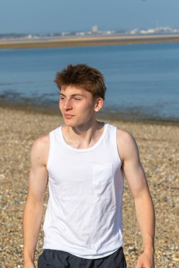 Beyaz şort ve kolsuz bluz giyen genç bir çocuk deniz kenarındaki taştan bir plajda ileri doğru yürüyor.