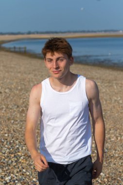 Beyaz şort ve kolsuz bluz giyen genç bir çocuk, deniz kıyısındaki taştan bir plajda ileri doğru yürüyor.