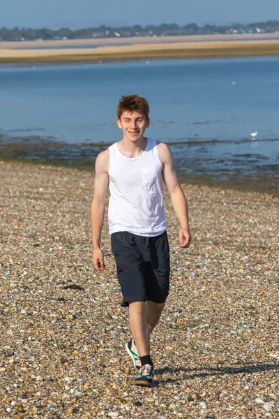 Beyaz şort ve kolsuz bluz giyen, deniz kıyısındaki taştan bir plajda yürüyen, gülümseyen genç bir çocuk.