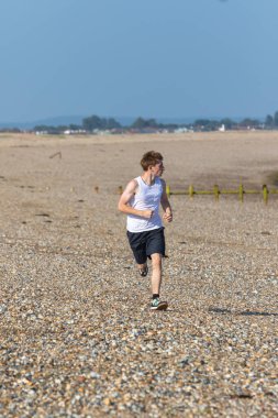 Genç bir çocuk sıcak bir yaz gününde taşlı bir plajda koşuyor ya da koşuyor. Egzersiz ve sağlıklı yaşam tarzı. Kameraya doğru gidiyor.