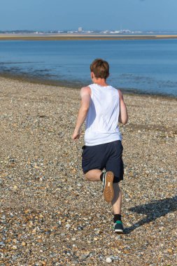 Genç bir çocuk sıcak bir yaz gününde taşlı bir plajda koşuyor ya da koşuyor. Egzersiz ve sağlıklı yaşam tarzı. Kameraya doğru gidiyor.
