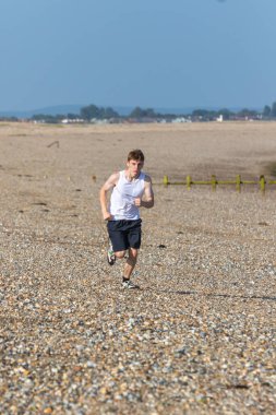 Genç bir çocuk sıcak bir yaz gününde taşlı bir plajda koşuyor ya da koşuyor. Egzersiz ve sağlıklı yaşam tarzı. Kameraya doğru gidiyor.