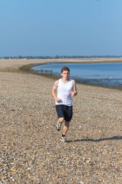 Genç bir çocuk sıcak bir yaz gününde taşlı bir plajda koşuyor ya da koşuyor. Egzersiz ve sağlıklı yaşam tarzı. Kameraya doğru gidiyor.