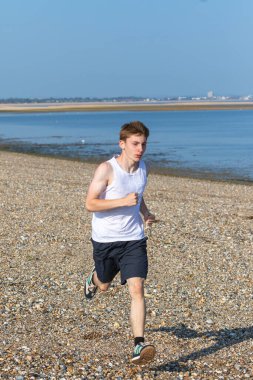 Genç bir çocuk sıcak bir yaz gününde taşlı bir plajda koşuyor ya da koşuyor. Egzersiz ve sağlıklı yaşam tarzı. Kameraya doğru gidiyor.
