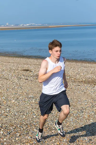 Genç bir çocuk sıcak bir yaz gününde taşlı bir plajda koşuyor ya da koşuyor. Egzersiz ve sağlıklı yaşam tarzı. Kameraya doğru gidiyor.