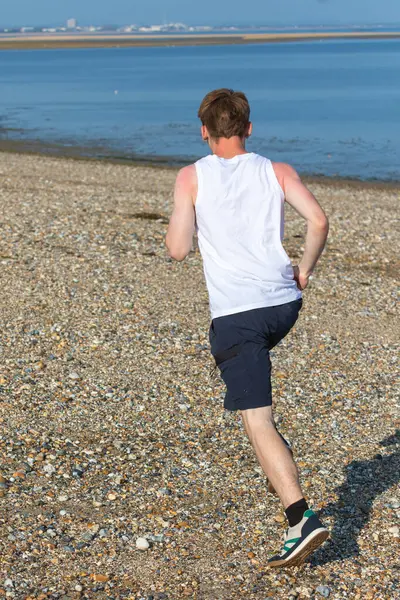 Genç bir çocuk sıcak bir yaz gününde taşlı bir plajda koşuyor ya da koşuyor. Egzersiz ve sağlıklı yaşam tarzı. Kameraya doğru gidiyor.