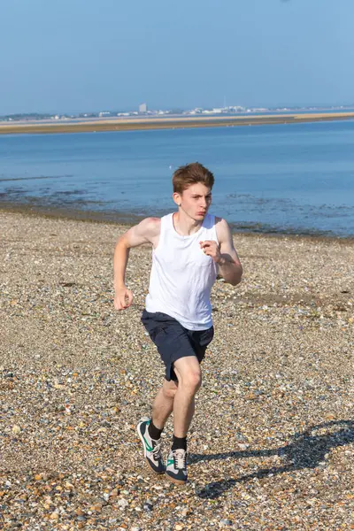 Genç bir çocuk sıcak bir yaz gününde taşlı bir plajda koşuyor ya da koşuyor. Egzersiz ve sağlıklı yaşam tarzı. Kameraya doğru gidiyor.