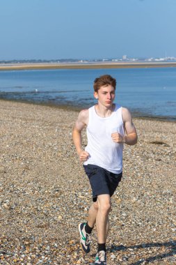 Genç bir çocuk sıcak bir yaz gününde taşlı bir plajda koşuyor ya da koşuyor. Egzersiz ve sağlıklı yaşam tarzı. Kameraya doğru gidiyor.