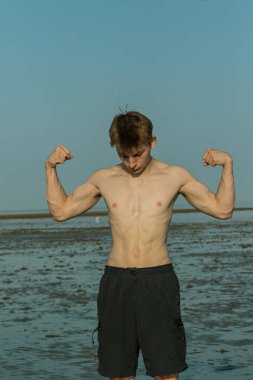 Tişörtsüz, kaslı ergen çocuk sıcak bir yaz gününde kumsalda kaslarını esnetiyor.