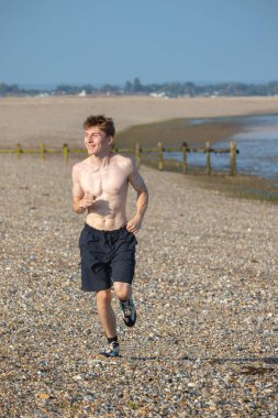 Tişörtsüz genç bir çocuk, sıcak bir yaz gününde, kayalık bir plajda kameraya doğru koşuyor.