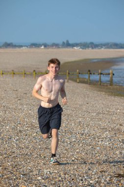 Tişörtsüz genç bir çocuk, sıcak bir yaz gününde, kayalık bir plajda kameraya doğru koşuyor.