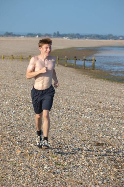 Tişörtsüz genç bir çocuk, sıcak bir yaz gününde, kayalık bir plajda kameraya doğru koşuyor.