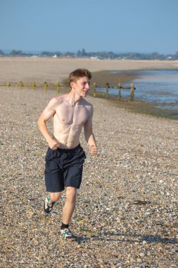 Tişörtsüz genç bir çocuk, sıcak bir yaz gününde, kayalık bir plajda kameraya doğru koşuyor.