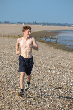 Tişörtsüz genç bir çocuk, sıcak bir yaz gününde, kayalık bir plajda kameraya doğru koşuyor.