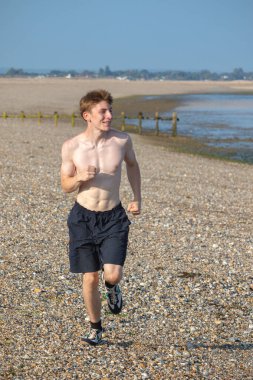 Tişörtsüz genç bir çocuk, sıcak bir yaz gününde, kayalık bir plajda kameraya doğru koşuyor.