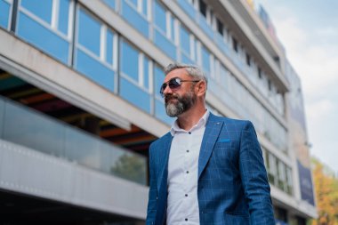 Yakışıklı, şık, olgun, güneş gözlüğü takan ve şehir binasının yakınında yürürken beyaz gömlek giyen bir adam. h