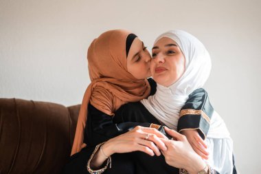 Mutlu Müslüman aile evlerinde küçük anne ve kız tesettürde öpüşür ve kanepede sevgiyle kucaklaşır anneler gününde birbirlerine bağlanmak için birlikte güler ve oynarlar. Yüksek kalite fotoğraf