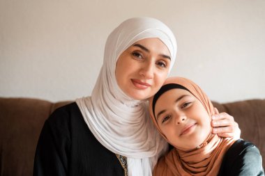 Evde mutlu bir Müslüman ailenin portresi tesettürlü genç anne ve kız kameraya bakıyor ve gülümsüyor. Yüksek kalite fotoğraf