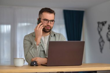 Genç bir adam ev ofisinde çalışırken telefonla konuşuyor ve dizüstü bilgisayar kullanıyor. Yüksek kalite fotoğraf