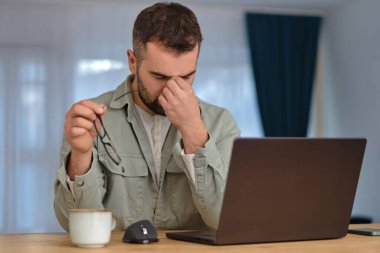 Fazla mesai yapan yorgun bir adam, dizüstü bilgisayarın önünde oturuyor, elinde gözlük tutuyor ve elleriyle gözlerini ovuşturuyor. Yüksek kalite fotoğraf