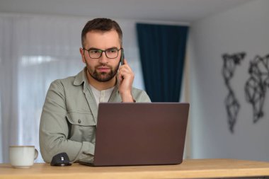 Genç bir adam ev ofisinde çalışırken telefonla konuşuyor ve dizüstü bilgisayar kullanıyor. Yüksek kalite fotoğraf