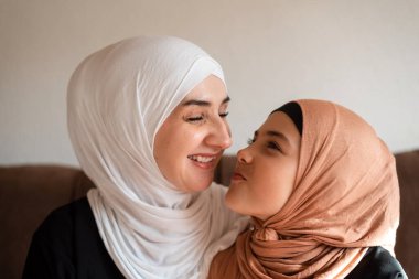 Mutlu Müslüman aile evlerinde küçük anne ve kız tesettürde öpüşür ve kanepede sevgiyle kucaklaşır anneler gününde birbirlerine bağlanmak için birlikte güler ve oynarlar. Yüksek kalite fotoğraf