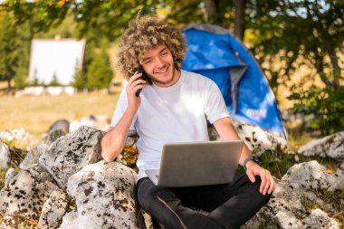 Telefonla konuşan ve dışarıda dizüstü bilgisayar kullanan genç ve mutlu bir adam. Yüksek kalite fotoğraf