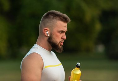 Genç ve zinde bir adam kablosuz kulaklıkla müzik dinliyor ve içme suyu içiyor ya da yaz parkında spor yaptıktan sonra biberondan beslenme suyu içiyor, aktif ve sağlıklı bir yaşam tarzı. Yüksek kalite fotoğraf